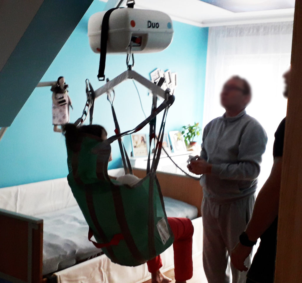 Osoba w podnośniku w pokoju, wspierana przez opiekunów.